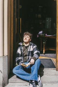Full length of young man sitting at entrance of bookstore on sunny day