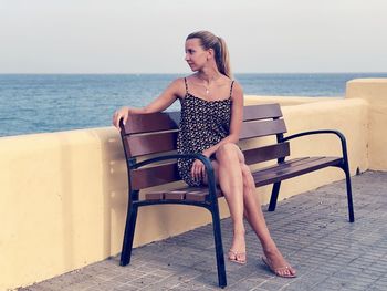 Young woman sitting on chair by sea against sky