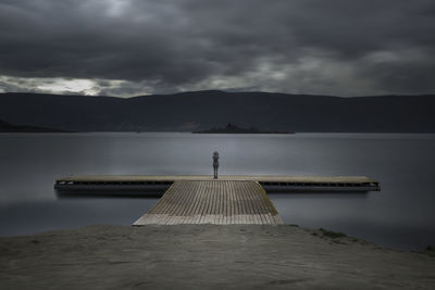 Mid distance view of woman standing on pier by lake against sky