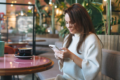 Attractive young brunette woman in white casual dress with cup of coffee using mobile phone in cafe