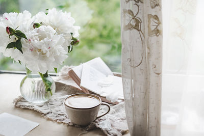 Stylish ceramic cup with  coffee and foam at window with curtains and glass vase peonies, cozy home