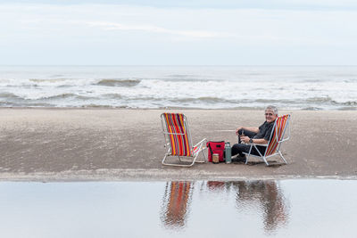 Mature man with gray hair sitting on a beach chair smiling while looking at the camera.