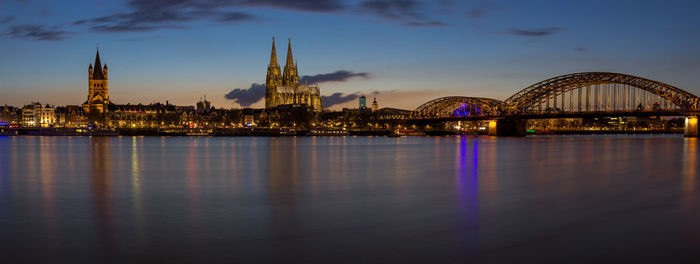 Illuminated bridge over river by buildings against sky in city