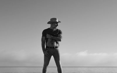 Monochrome of adult man in cowboy hat taking off cloth on beach against sky