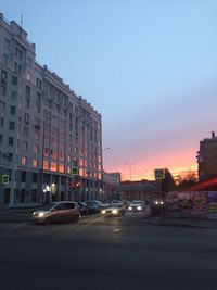 Cars on road by buildings against sky at sunset