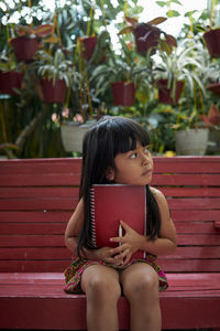 Girl with book looking away while sitting on bench