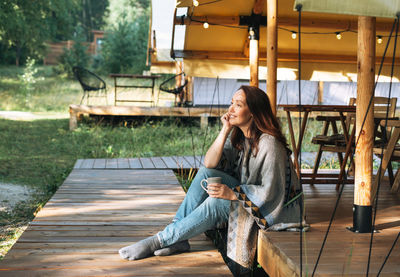 Young brunette woman in poncho drinking tea and relaxing in glamping in nature