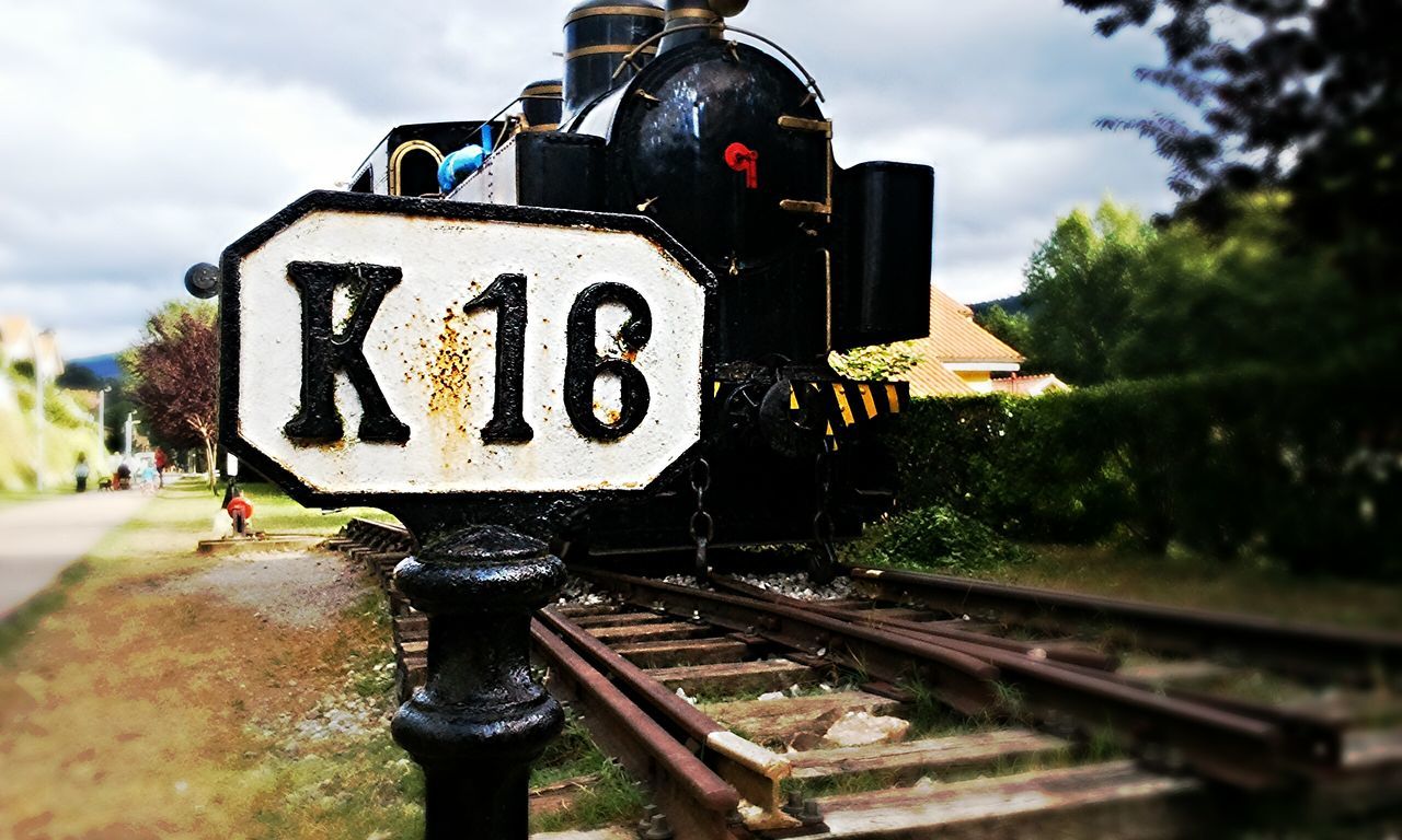 text, communication, sky, western script, transportation, railroad track, cloud - sky, road sign, focus on foreground, information sign, tree, sign, rail transportation, day, outdoors, guidance, close-up, mode of transport, land vehicle, warning sign