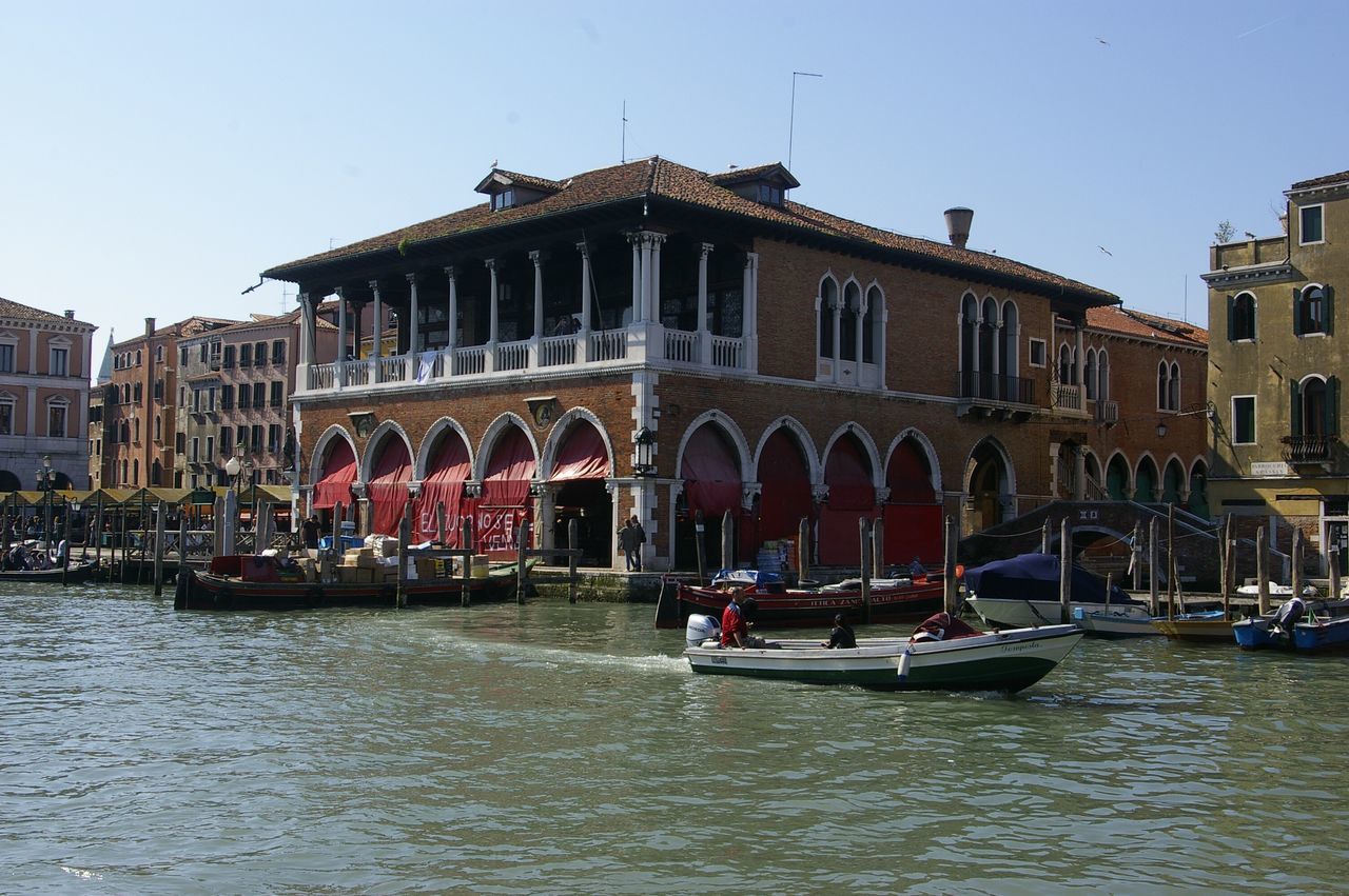 building exterior, built structure, architecture, water, waterfront, nautical vessel, transportation, boat, window, mode of transport, city, arch, canal, travel destinations, clear sky, city life, facade, arcade, in front of, river, old town, day, gondola - traditional boat, tourism, town square, outdoors, culture, famous place, sky