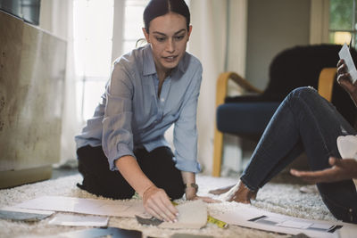 Female designer discussing about fabric swatch with colleague while kneeling on carpet at home office