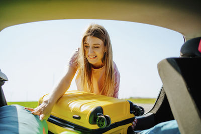 Happy girl loading yellow suitcase in car trunk