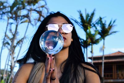 Woman wearing sunglasses holding crystal ball against palm trees