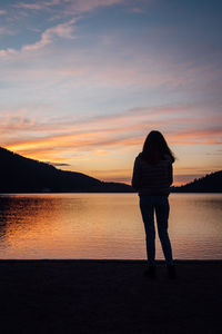 Rear view of silhouette girl standing by lake against sky during sunset