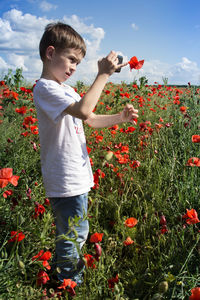 Boy standing by flowers on field against sky