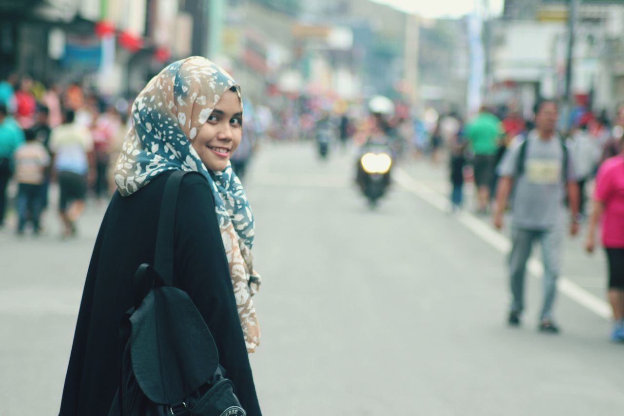 focus on foreground, lifestyles, leisure activity, front view, young adult, portrait, street, casual clothing, looking at camera, warm clothing, person, incidental people, winter, standing, building exterior, smiling, city, waist up