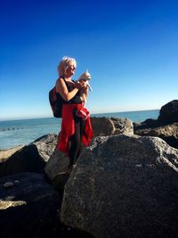 Woman holding dog while standing on rocky shore against clear blue sky