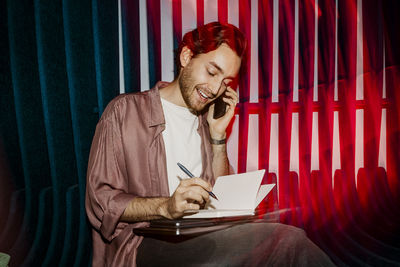 Smiling male entrepreneur talking on smart phone and writing in diary while sitting on seat