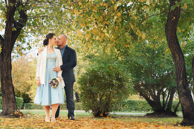 Bride and groom embracing while standing by tree trunk