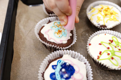 A child squeezes colored frosting from a tube onto chocolate brown cupcakes covered white frosting.