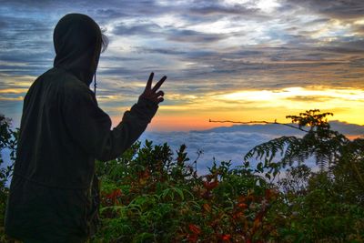 Rear view of woman gesturing peace sign while standing by plants against sky during sunset