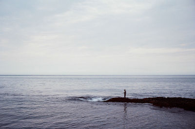 Man standing on rock formation in sea while fishing against sky