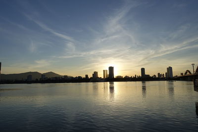 Silhouette buildings by river against sky during sunset