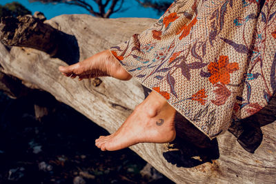 Unrecognizable crop woman in summer dress and with tattoo on leg sitting on tree trunk in forest