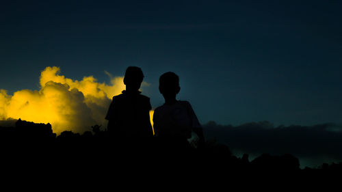 Silhouette siblings standing against sky at sunset