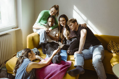 Young woman sharing smart phone with multiracial friends sitting together on sofa at home