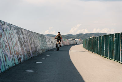 Low angle of confident young female bicyclist in sportswear and helmet riding bike on fenced curvy paved track while training alone in summer day