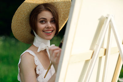 Portrait of smiling young woman wearing hat standing against wall