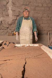 Calm peruvian male worker with dirty hands in uniform looking at camera while standing near plaster near wall during work in ceramics workshop