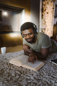 Smiling man reading book while listening to music through headphones lying on carpet at home