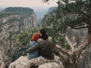 Rear view of couple sitting on cliff against mountains 