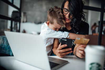 Boy embracing mother using phone while holding credit card