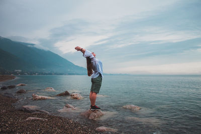 Side view of woman stretching while standing on rock by sea against cloudy sky