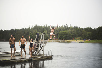 Male and female friends enjoying on jetty over lake against clear sky during weekend getaway