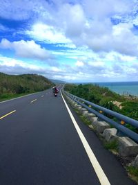 Empty road along calm sea against cloudy sky