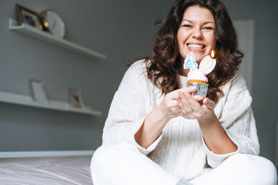 Young adult woman forty years plus size body positive with cupcake with candle in hands at home 
