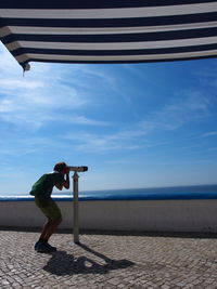 Man looking through coin-operated binoculars by sea against sky