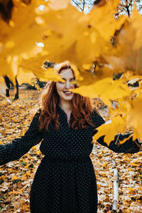 Happy red-haired woman holding yellow maple autumn leaves in fall park.