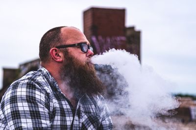 Man exhaling smoke while sitting against sky