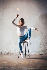 Portrait of woman holding lit sparkler while sitting on table against wall