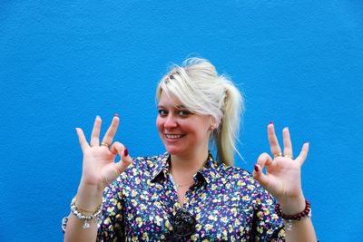 Portrait of smiling young woman gesturing ok sign against blue wall