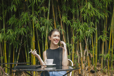 Smiling woman talking over smart phone and smoking cigarette while sitting at outdoor cafe