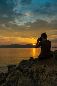 Man photographing rock on sea against sky during sunset