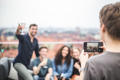 Rear view of woman photographing friends through mobile phone during terrace party