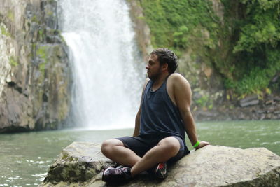 Young man sitting on rock against waterfall