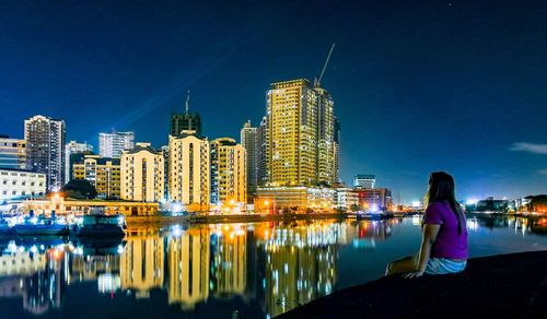 Woman sitting at riverbank against illuminated buildings in city