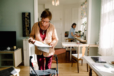Senior woman reading newspaper while caregiver working in kitchen at nursing home